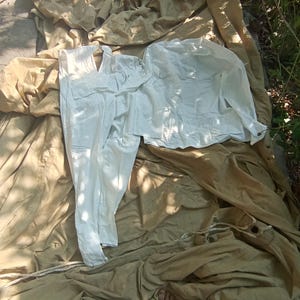 Könnte beinhalten: Ein weißes Langarmhemd und eine passende weiße Hose, die auf einem großen, beigefarbenen Stofftuch ausgebreitet sind. Das Hemd hat einen Kragen und eine durchgehende Knopfleiste, während die Hose einen geraden Schnitt aufweist. Der Stoff wirkt leicht.