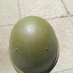 May include: An olive green military-style helmet with a smooth, rounded dome shape. The helmet features a wide, slightly flared brim and small, evenly spaced metal rivets around the lower perimeter, showing a weathered, matte finish.