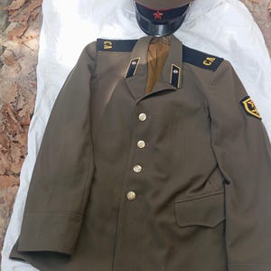 May include: An olive green military uniform jacket featuring gold-tone buttons, black shoulder boards with the letters CA, and a matching peaked cap with a red star emblem. The jacket includes dark collar tabs with white star insignias.