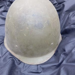 Puede incluir: Un casco militar verde oliva con arañazos y desgaste visibles. El casco tiene una parte superior redondeada y un borde ligeramente ensanchado. Descansa sobre una superficie oscura y texturizada, lo que sugiere un estado vintage o usado.