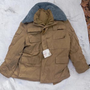 May include: A tan field jacket featuring a gray faux fur collar, four large front pockets with flaps, and a small white tag attached to the center button. The jacket has long sleeves and a structured design with visible stitching throughout.