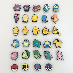 Könnte beinhalten: Eine Sammlung farbenfroher Schuhanhänger mit Cartoon-Charakteren. Die Anhänger zeigen verschiedene Designs, darunter beliebte Charaktere und Pokéball-Designs. Die Anhänger bestehen aus glänzendem Material und sind auf einer weißen Oberfläche angeordnet.