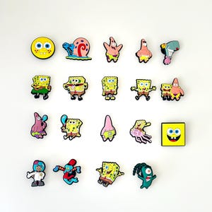 Könnte beinhalten: Eine Sammlung farbenfroher Cartoon-Charakter-Anhänger, darunter SpongeBob Schwammkopf, Patrick Star und andere Charaktere. Die Anhänger sind in verschiedenen Posen und Designs gehalten, mit leuchtenden Gelb-, Rosa- und Grüntönen. Sie sind wahrscheinlich für Schuhe oder Accessoires.