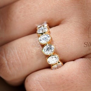 May include: A gold band ring featuring a series of oval-cut clear stones set in a bezel style. The ring displays the text THE SWARNA ELEGANCE with a small diamond icon logo printed on the side. The stones are arranged in a continuous row.
