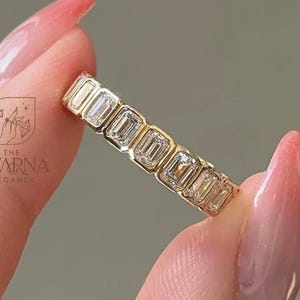 以下が含まれることがあります： 個別のベゼルにセットされた7つのエメラルドカットの透明な石が並ぶゴールドのバンドリング。リングには「THE SWARNA ELEGANCE」というテキストと、ダイヤモンドを持つ手のアイコンが描かれたロゴが表示されています。