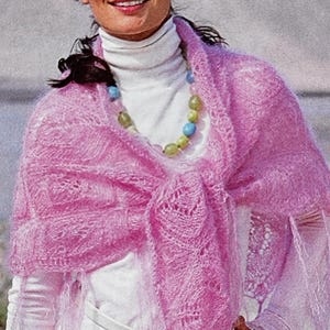 May include: A woman wearing a light pink, open-weave shawl over a white turtleneck and white outfit. The shawl has a fringed edge and is draped around the shoulders. A beaded necklace with blue and green beads is visible.
