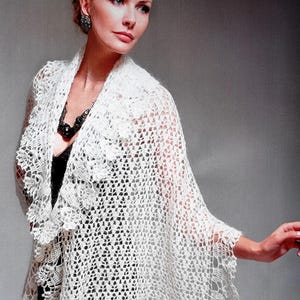 May include: A white, open-weave crochet shawl with a scalloped edge. The shawl is draped over a black garment. The image is set against a neutral background, highlighting the intricate lace-like design of the shawl.