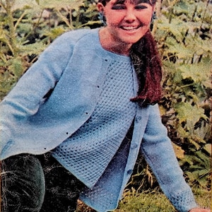 May include: A light blue cardigan and sweater set. The cardigan has a button-up front and long sleeves. The sweater has a textured, open-knit design. The person is sitting on the grass in front of a green leafy background.