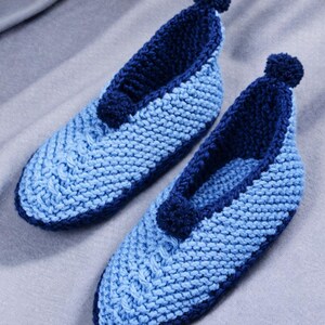 May include: A pair of knitted slippers in shades of blue. The slippers have a light blue body with a darker blue trim and a small pom-pom detail. The knitted pattern is visible. The slippers are made by hand.