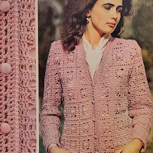 May include: A pale pink, open-weave crochet cardigan with long sleeves and a V-neck. The cardigan has a button-up front with large, round buttons. The crochet pattern features floral and openwork designs. The garment is worn over a white blouse.