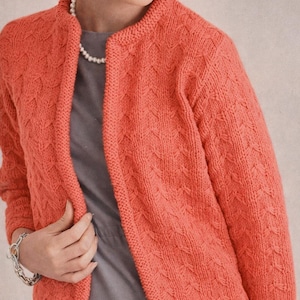 May include: Coral-colored knitted cardigan with a textured pattern. The open-front sweater has a round neckline and long sleeves. The cardigan is worn over a gray dress. A silver bracelet and a pearl necklace are also visible.