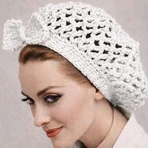 May include: A white, open-weave crochet beret with a decorative bow on the side. The hat is made of a thick, textured yarn and is worn on the head. The beret is a classic accessory.
