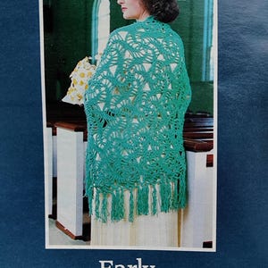 May include: A teal crocheted shawl with fringe, worn by a person wearing a white hat. The shawl has an open, lacy design. The text "Early Spring Shawl" is at the bottom of the image. The background shows a church interior.