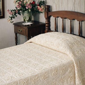 Puede incluir: Una colcha de ganchillo de color crema con un patrón floral y flecos. La colcha cubre una cama con cabecero de madera. Un jarrón con flores está sobre una mesita de noche de madera.