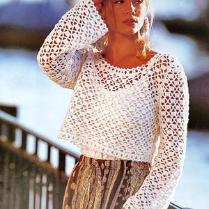 Può includere: Un top all'uncinetto bianco a trama aperta con maniche lunghe. Il top è abbinato a una gonna fantasia. Il design all'uncinetto presenta un motivo geometrico. L'immagine è ambientata all'aperto con uno sfondo sfocato.