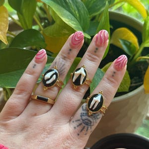 May include: A hand displays four gold rings, three with black oval settings featuring a white tooth design. The fourth ring is a simple gold band with a black rectangular inlay. The nails are painted with pink and white swirls.