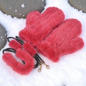 Può includere: Un paio di guanti in pelliccia rosso vivo e una fascia abbinata poggiano su una superficie innevata. I guanti hanno un design classico e la fascia è collegata ai guanti con un cordino nero. I fermagli color oro fissano il cordino.