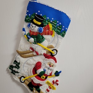 May include: A Christmas stocking with a blue top adorned with sequins. The stocking features a snowman and Santa Claus skiing down a snowy hill. The snowman wears a black hat, earmuffs, and a red scarf. Santa wears a red suit and hat. The words "North Pole" are on a yellow sign.