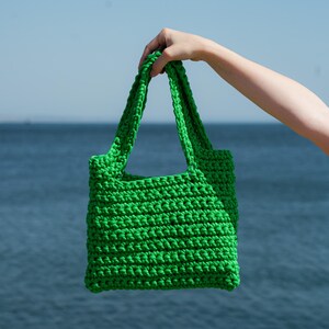 May include: A bright green, hand-crocheted tote bag with sturdy handles. The bag is held up against a backdrop of a blue ocean and sky. The bag's texture is visible, showcasing the tight stitches of the crochet.