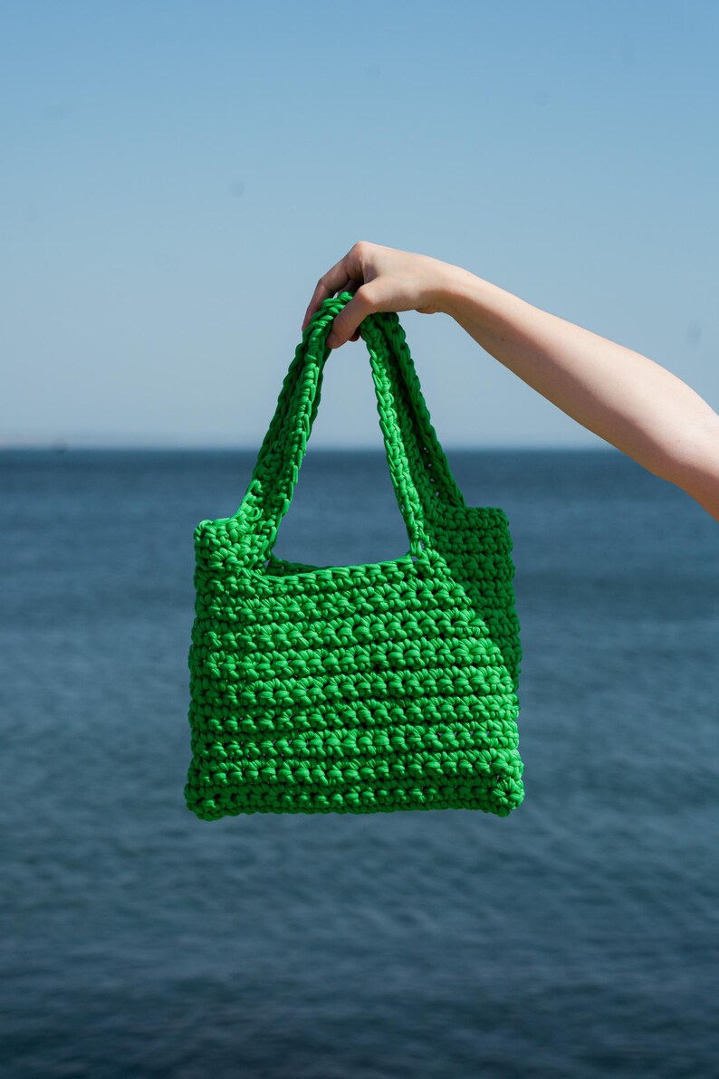 May include: A bright green, hand-crocheted tote bag with sturdy handles. The bag is held up against a backdrop of a blue ocean and sky. The bag's texture is visible, showcasing the tight stitches of the crochet.