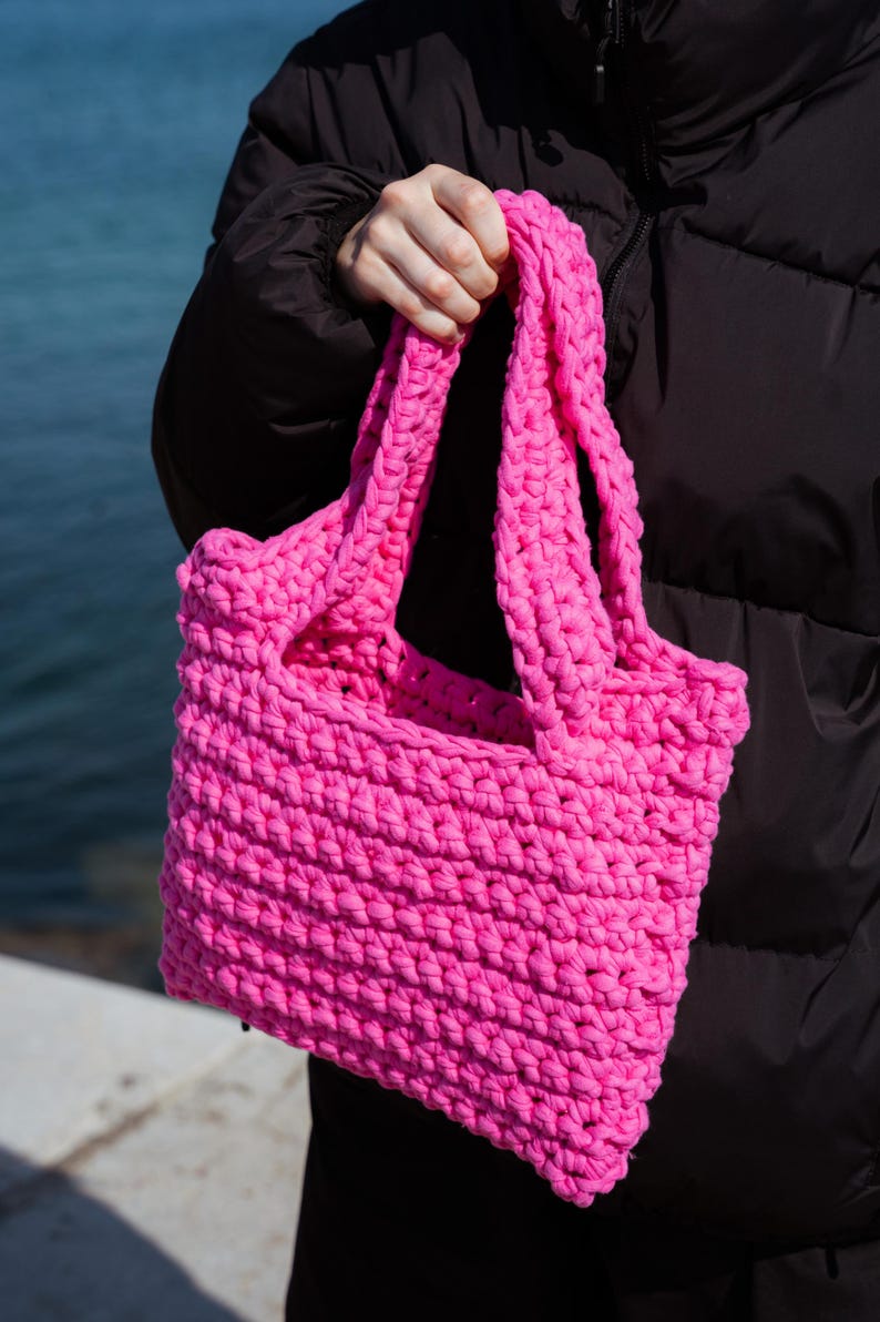 May include: A bright pink, hand-crocheted tote bag. The bag is rectangular with two sturdy handles. The thick yarn creates a textured surface. The bag is held against a dark jacket and a blue water background.