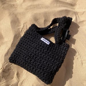 May include: A black crocheted tote bag with short handles and a rectangular shape. The bag has a small white tag with black text. The bag is sitting on a sandy surface.