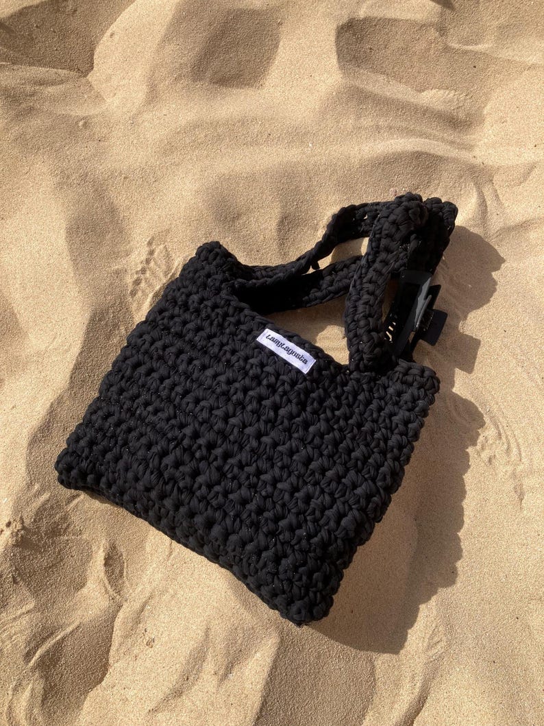 May include: A black crocheted tote bag with short handles and a rectangular shape. The bag has a small white tag with black text. The bag is sitting on a sandy surface.