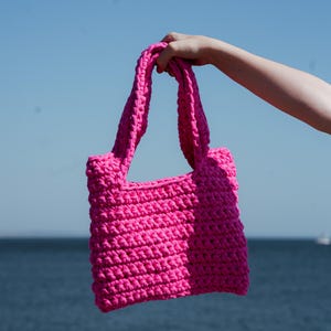 May include: A bright pink, hand-crocheted tote bag with sturdy handles. The bag is held up against a backdrop of a blue sky and ocean. The bag's texture is visible, showcasing the tight stitches.