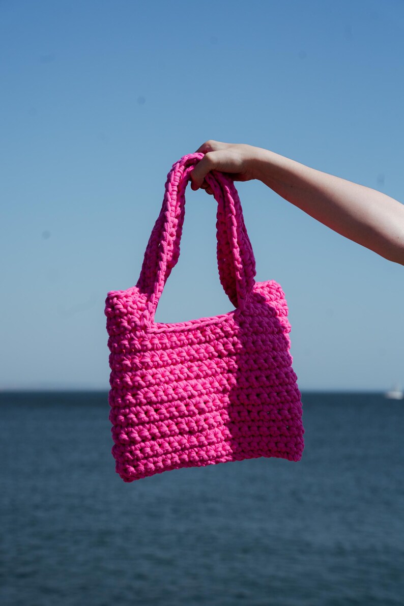 May include: A bright pink, hand-crocheted tote bag with sturdy handles. The bag is held up against a backdrop of a blue sky and ocean. The bag's texture is visible, showcasing the tight stitches.