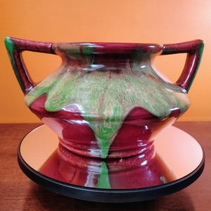 May include: A maroon ceramic vase with two handles, featuring a green and brown glaze that drips down the sides. The vase sits on a reflective surface, showcasing its glossy finish. The overall design is vintage and decorative.