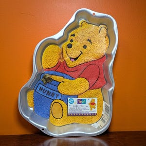 May include: A silver-colored metal cake pan shaped like Winnie the Pooh holding a blue honey pot with the word HUNNY printed on it. The pan features a textured interior surface and includes a product label with the text Pooh Cake Pan.