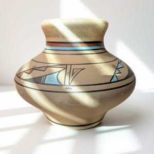 May include: A tan ceramic vase with a wide, rounded body and a flared neck. The surface features hand-painted geometric patterns in black, light blue, and terracotta, with the signature MN02 visible on the lower section of the vessel.