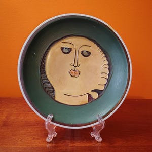 May include: A ceramic plate with a face design. The plate features a green rim and a beige face with dark eyes and a pursed mouth. The face is surrounded by a textured border. The plate is displayed on a clear stand.
