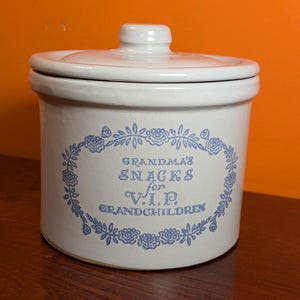 May include: A cylindrical off-white ceramic jar with a matching lid. The front features a blue floral wreath design surrounding the text: GRANDMA'S SNACKS for V.I.P. GRANDCHILDREN. The jar has a smooth finish and a simple knob on the lid.