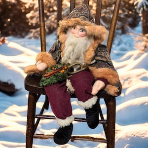 May include: A fabric doll with a long white beard, wearing a patterned hood and cloak with faux fur trim, and dark red trousers. The doll holds a small green wreath with orange berries and a brass-colored trumpet while sitting on a wooden chair.