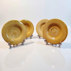 May include: A set of four circular ceramic saucers in a warm mustard yellow glaze. Each saucer features a smooth, slightly concave surface with a central indentation designed to hold a cup, displayed upright on small silver-toned metal stands.