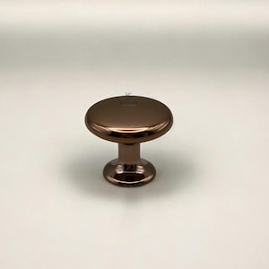 May include: A bronze-colored cabinet knob with a round, flat top and a short, cylindrical base. The knob has a shiny, reflective surface and is set against a plain white background. The word "KULFERA" is printed on the top of the knob.