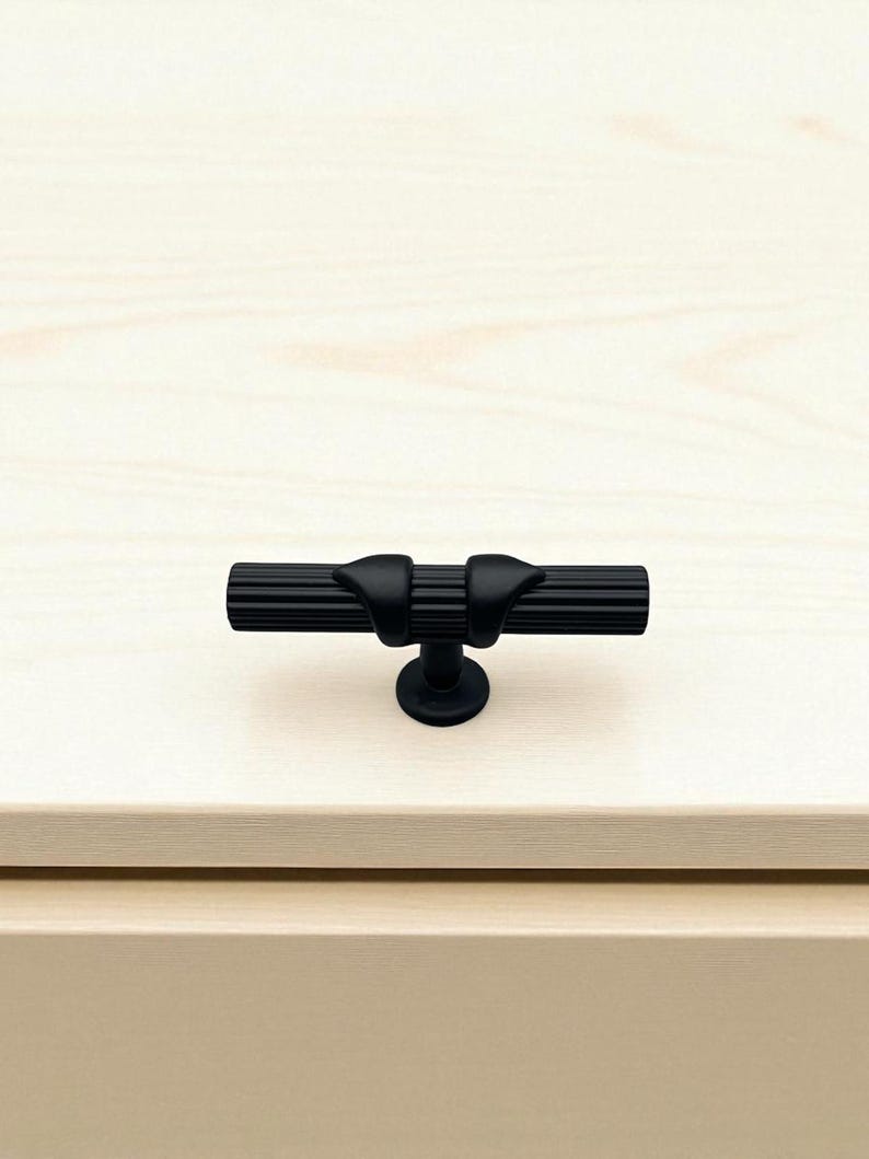Może przedstawiać: Czarny uchwyt do szafki z cylindrycznym uchwytem i dwoma zakrzywionymi akcentami. Uchwyt jest zamontowany na małej, okrągłej podstawie. Okucia są osadzone na jasnej drewnianej powierzchni.