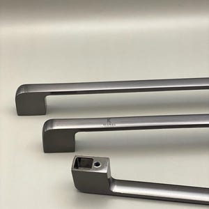 Puede incluir: Tres tiradores de armario de metal cepillado de diferentes tamaños. Los tiradores son de color gris apagado con un diseño rectangular y bordes redondeados. El nombre de la marca "KUPERA" está impreso en uno de los tiradores.