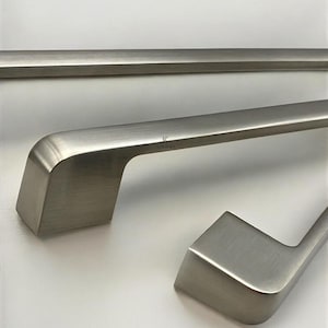 May include: Brushed nickel cabinet hardware. The image shows three pieces of cabinet hardware with a brushed nickel finish. The hardware has a modern design with a rectangular base and a curved handle. The hardware is designed for cabinets and drawers.