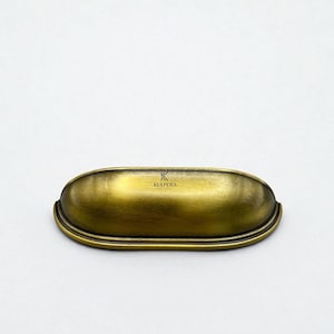 Puede incluir: Un tirador de cajón de estilo vintage, en tono latón, con un diseño ovalado y curvado. El herraje presenta un acabado ligeramente envejecido, lo que añade a su estética antigua. La marca "KULPERA" está visible en el tirador.