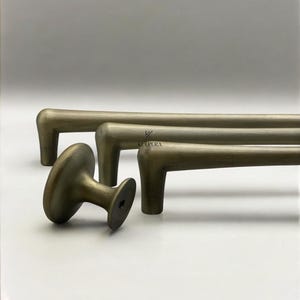 May include: Antique brass cabinet hardware set. The set includes two long bar pulls and a round knob. The hardware has a vintage finish and a classic design. The logo "KULPERA" is visible on the hardware.