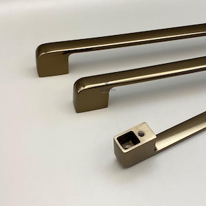 Könnte beinhalten: Drei bronzefarbene Schrankgriffe mit rechteckigem Design. Die Griffe haben eine glatte, metallische Oberfläche und sind auf weißem Hintergrund angeordnet. Die Griffe sind für eine einfache Installation konzipiert und verleihen jedem Schrank eine moderne Note.