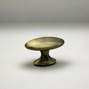 May include: An antique brass cabinet knob with an oval top and a curved base. The knob has a warm, aged finish and is designed for furniture. The brand name "Klipera" is subtly printed on the top.