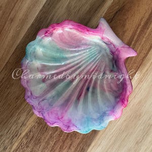 Op de afbeelding: Een schelpvormige sieradenschaal met een wervelend ontwerp in roze, blauw en wit. De schaal heeft een glanzende afwerking en de tekst "Charmed by Midnight" is zichtbaar.