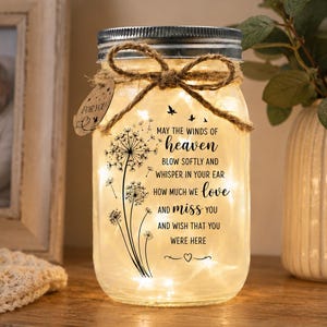 May include: A clear glass mason jar with a silver lid and a jute twine bow. The jar contains warm fairy lights and features a black dandelion graphic with the text: May the winds of heaven blow softly and whisper in your ear how much we love and miss you and wish that you were here. A small tag labeled For You is attached.