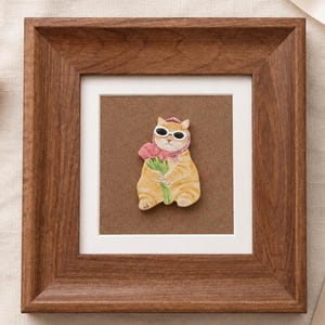 May include: A framed artwork features a whimsical cat illustration. The orange tabby cat wears sunglasses and a pink scarf, holding pink tulips. The artwork is set in a brown wooden frame with a white mat.