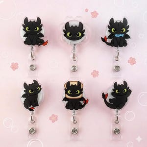 May include: Six retractable badge holders featuring a black cartoon dragon with green eyes and red accents. The dragon is in various poses, some with wings or accessories. The holders have a clear plastic base and a metal clip.