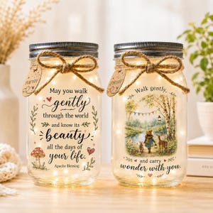 May include: Two clear glass mason jars with metal lids, each wrapped with a jute twine bow and a 'FOR YOU' tag. The jars contain warm fairy lights. One jar features the text 'May you walk gently through the world and know its beauty all the days of your life. Apache Blessing' with a mushroom illustration. The other jar shows a forest scene with a child, a deer, and a rabbit, with the text 'Walk gently, and carry wonder with you.'