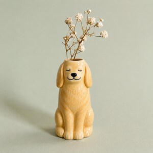 May include: A ceramic dog-shaped vase in a light yellow color, with closed eyes and a smiling expression. The vase holds dried white flowers. The dog vase is sitting upright on a light green background.